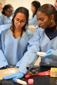 Two students wearing protective lab gowns and gloves work together during a hands-on pharmaceutical sciences lab activity at Campbell University.