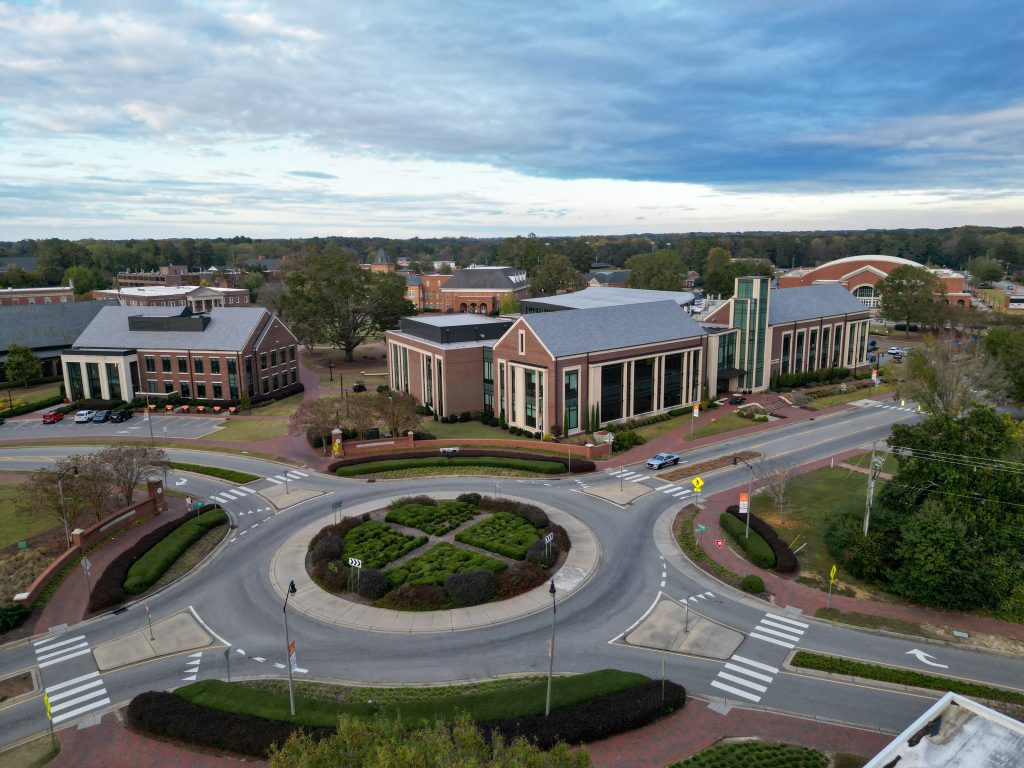 Aerial view of Campbell University's campus centered on the Oscar N. Harris Student Union and the campus roundabout. Brick academic buildings, tree-lined walkways and landscaped grounds surround the intersection, with the John W. Pope Jr. Convocation Center visible in the background beneath dramatic blue-gray clouds.