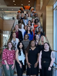 Students pose together on a wide staircase inside the Campbell University College of Pharmacy & Health Sciences building. The group is arranged on multiple steps and smiles for the camera in business and business-casual attire.