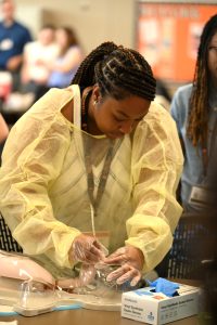 A student wearing protective gloves and a disposable gown practices airway management skills using a medical simulation mannequin.