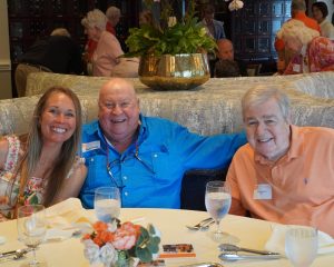Three attendees smile while seated together at a table during the Golden Club Luncheon, with other guests visiting in the background.