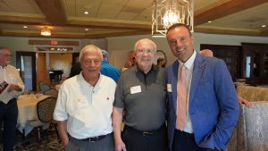 Three attendees smile while seated together at a table during the Golden Club Luncheon, with other guests visiting in the background.
