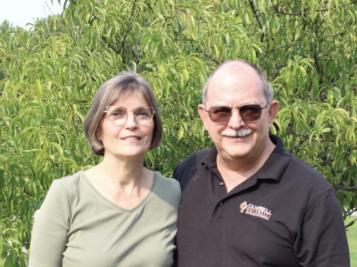 Two people are smiling outdoors in front of a leafy tree. One person is wearing glasses and a light green top. The other person, also wearing glasses, has a black "Campbell University" shirt.