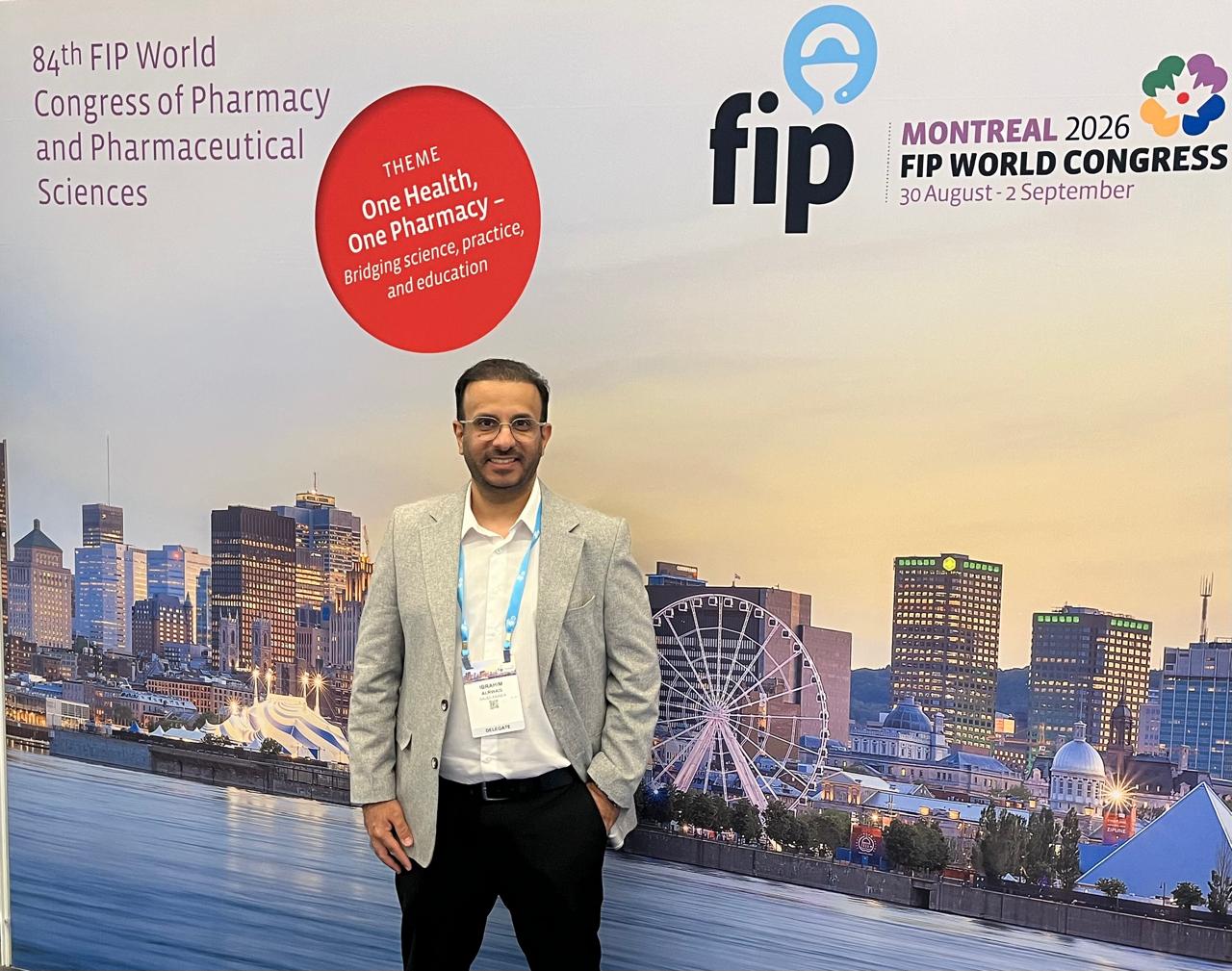 Man in grey suit coat and dark pants wearing lanyard standing in front of large conference banner