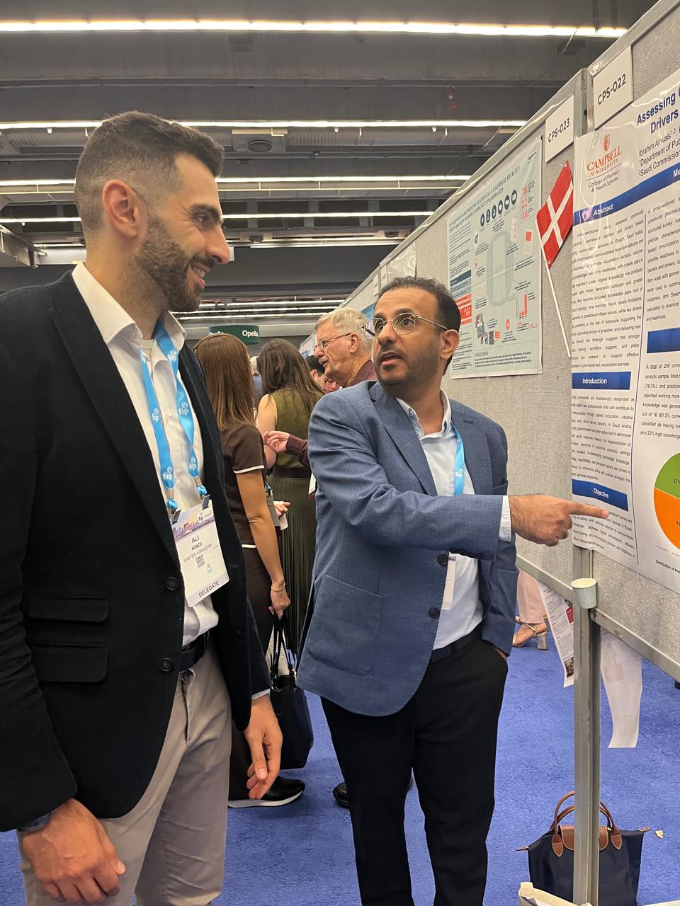 Man in blue suit coat and dark pants wearing lanyard standing in front of research poster talking with conference attendee