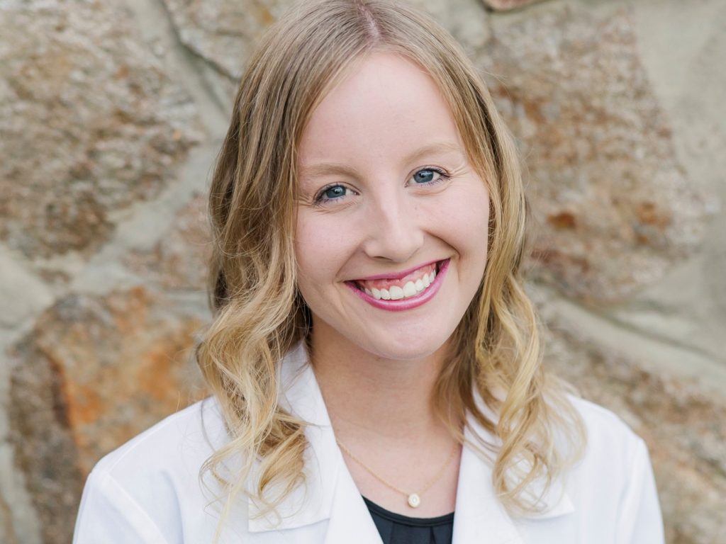 Blonde female student in a white lab coat with a big smile standing against a stone wall.