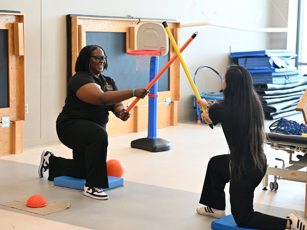 Two students kneeling on soft mats holding colorful weighted sticks and crossing them in the air