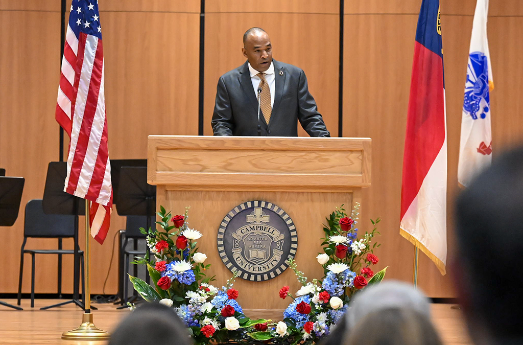 A person standing at a podium on a stage, speaking to an audience. The podium is surrounded by a floral arrangement and displays a seal. The backdrop includes the United States flag and other flags. The room is an auditorium with several rows of seats.