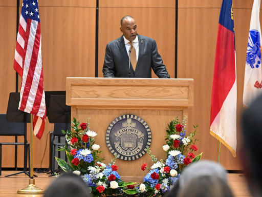 A person standing at a podium on a stage, speaking to an audience. The podium is surrounded by a floral arrangement and displays a seal. The backdrop includes the United States flag and other flags. The room is an auditorium with several rows of seats.