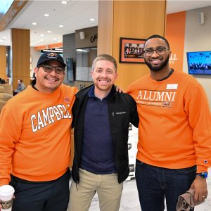 Three men pose with their arms around one another in the Student Union lobby, two wearing orange Campbell sweatshirts.