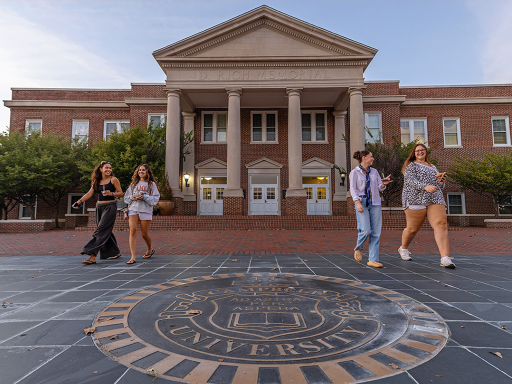 Students walk outside D. Rich Memorial Hall at Campbell University, with the University seal in the foreground.