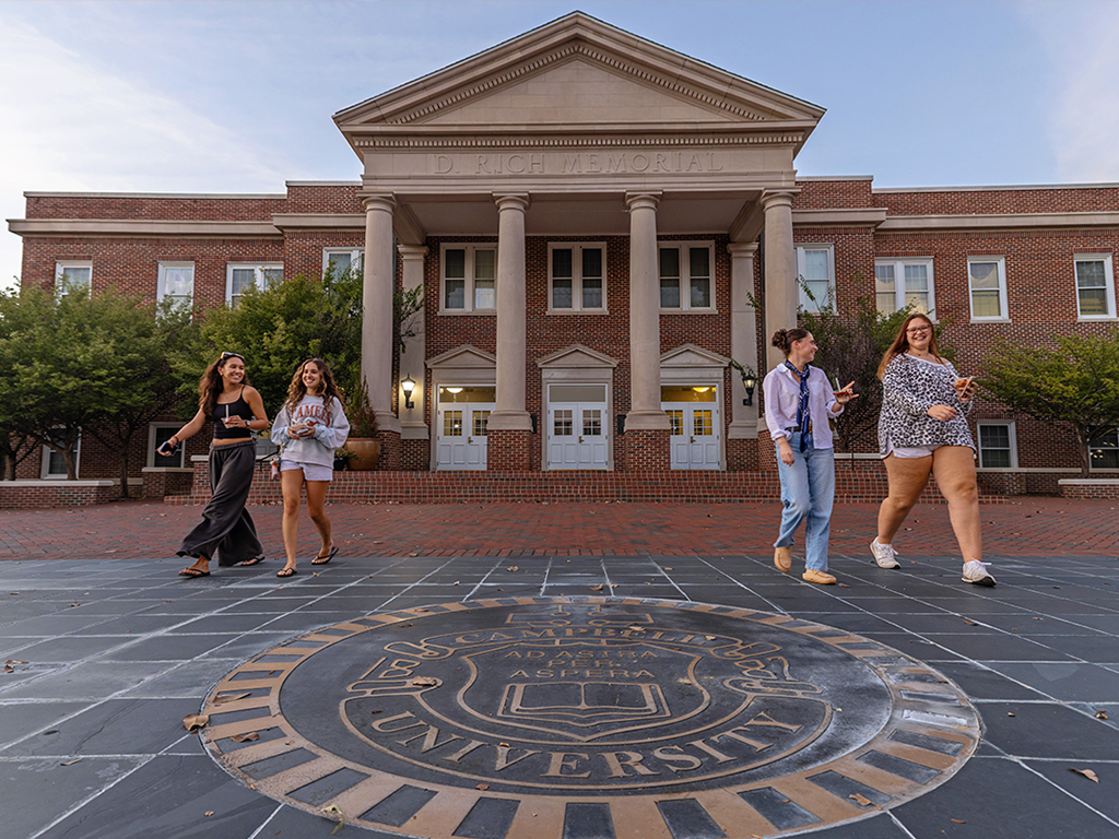 Students walk outside D. Rich Memorial Hall at Campbell University, with the University seal in the foreground.
