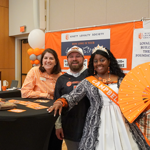 Three guests pose at the Kivett Loyalty Society booth, including a woman wearing a crown and Campbell sash and holding a large orange fan.