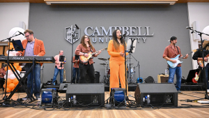 A singer in an orange jumpsuit performs with a live band onstage in the Keith Family Ballroom during the Alumni Block Party.