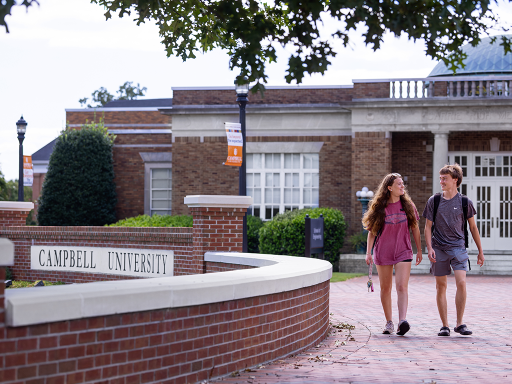 Two people are walking on a path in front of a building at Campbell University. The scene includes greenery and a clear sky.