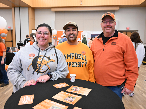 Three attendees wearing Campbell apparel smile together around a table at the 2026 Alumni Block Party in the Student Union.