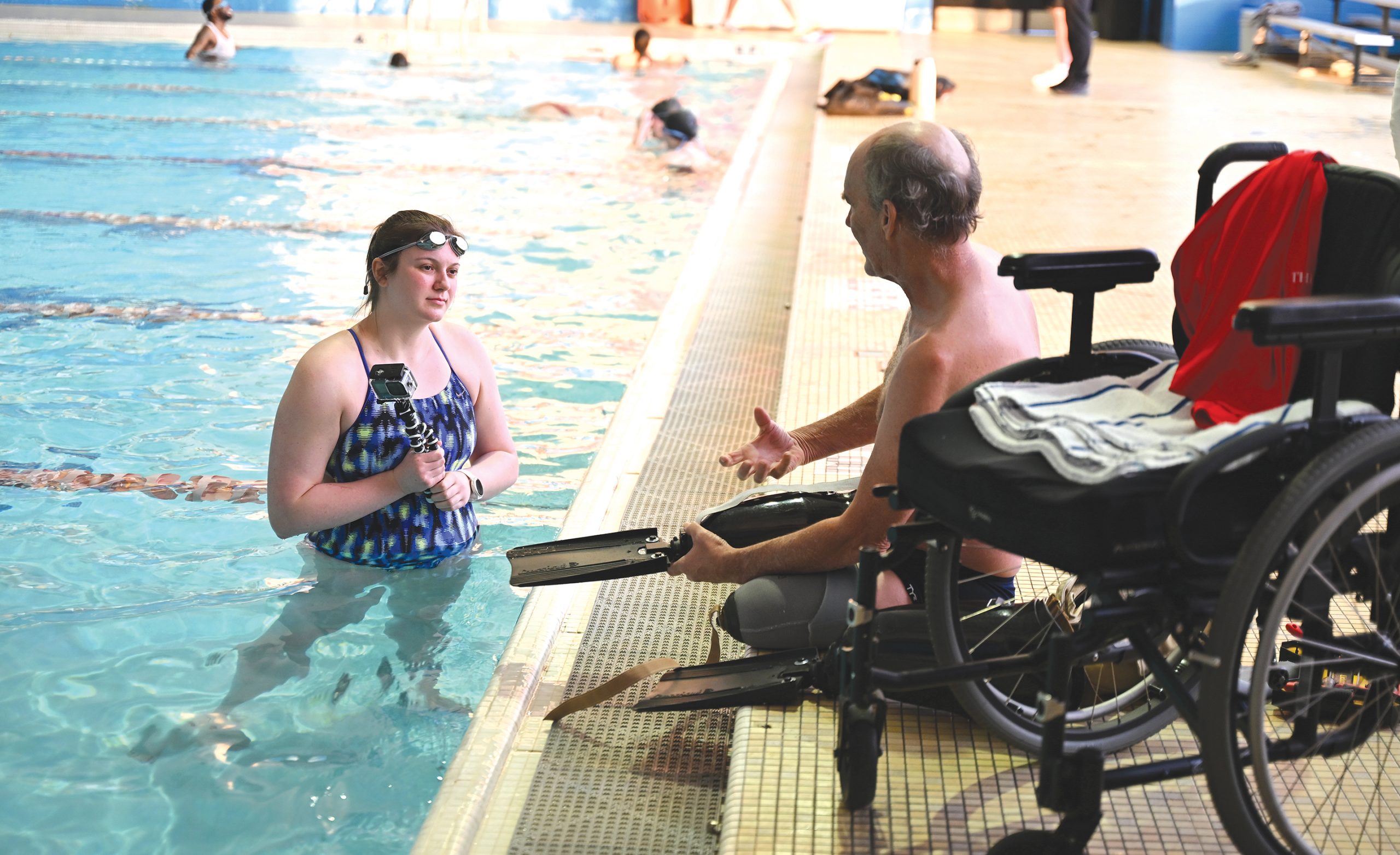 A student holds a camera in a swimming pool talking to a man as he tries on prosthetic swim fins.