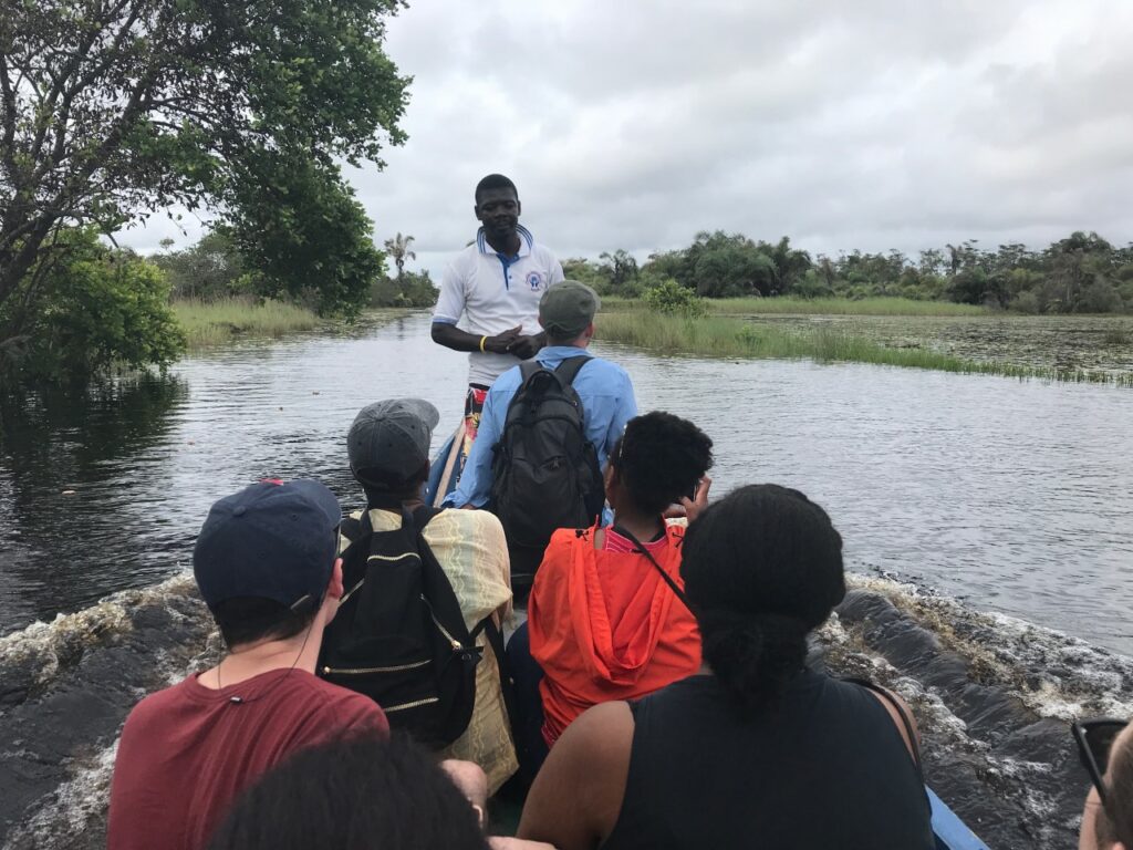 Photo of a five students backs seated in a wooden boat with a guide in a white shirt standing at the front as they glide down a river