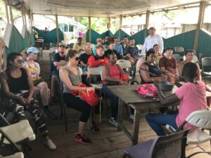 Photo of students, faculty and staff members sitting in front of a village member seated at a wooden table talking in an open-air room