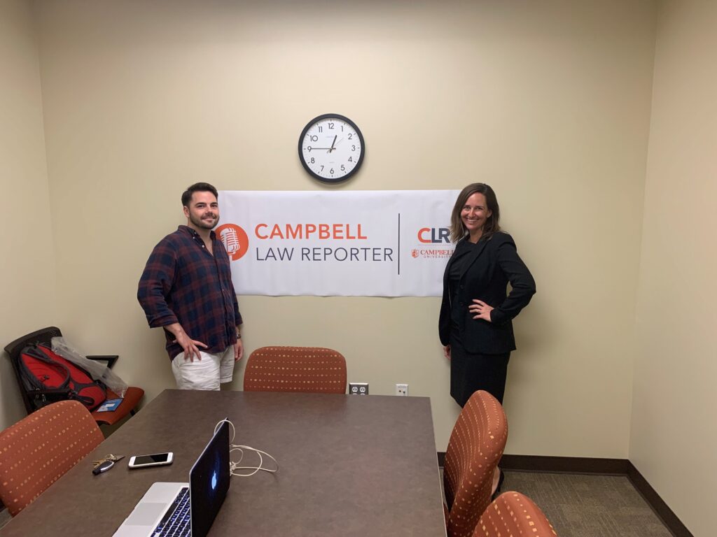 Photo of Bryant Pernell '20 and Professor Elizabeth Berenguer standing in frnot of the Campbell Law Reporter banner hanging in a confernce room under a wall clock