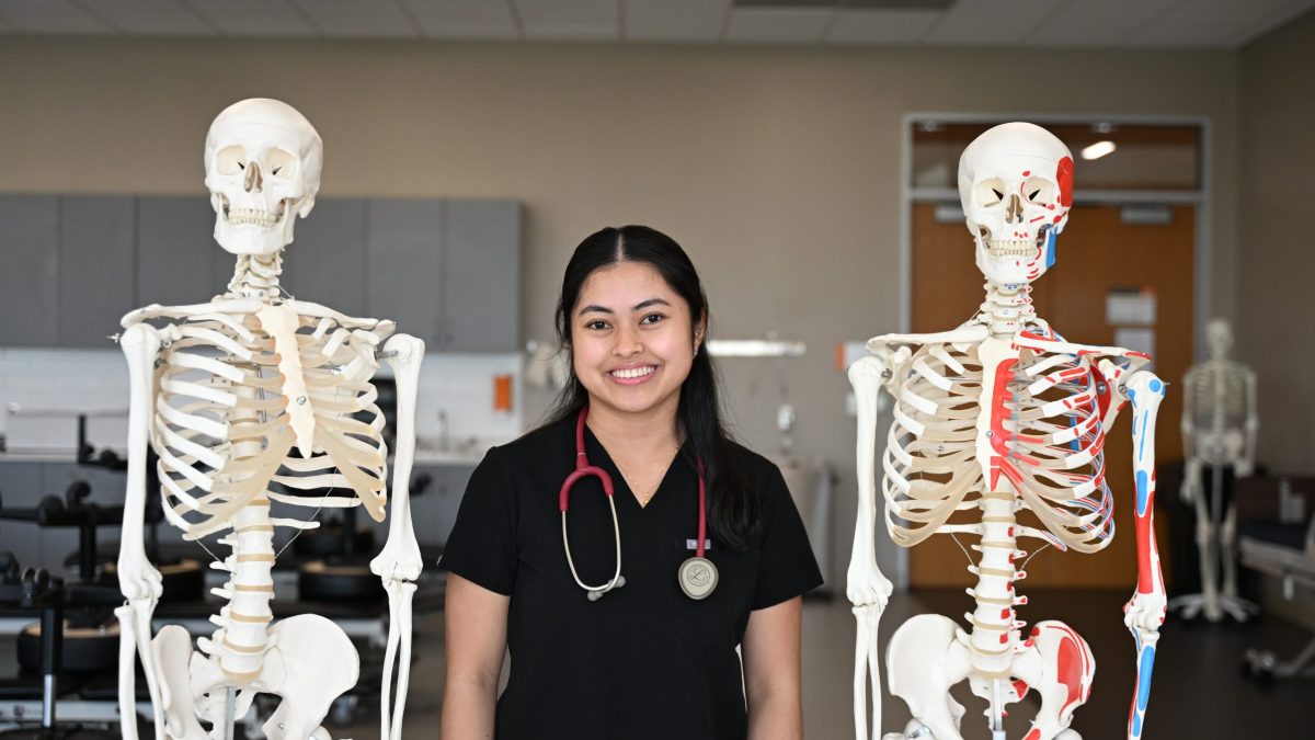 Female DPT student wearing black scrubs stands between two skeletons on stands