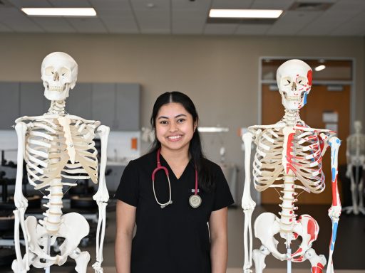 Female DPT student wearing black scrubs stands between two skeletons on stands