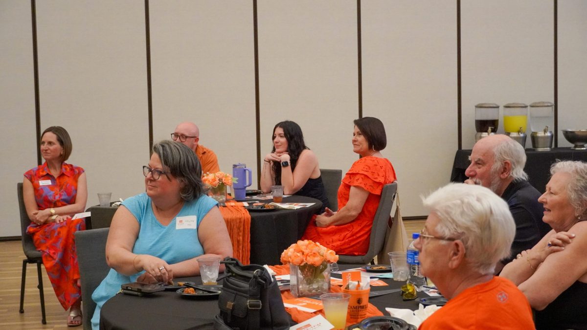 Two tables filled with people look attentively to the left. Many people are wearing orange. Orange flowers are on the tables.