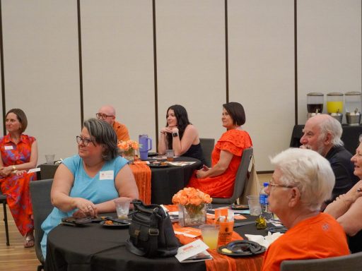 Two tables filled with people look attentively to the left. Many people are wearing orange. Orange flowers are on the tables.