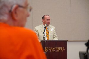 James Adcock ('09) speaking at a podium with Campbell University labeled on it. James is wearing a tan suit with orange and black striped tie.