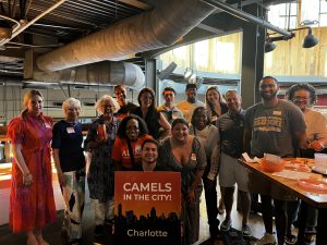 A group of people posing together indoors, holding a sign that reads "Camels in the City! Charlotte." They are standing in a casually decorated venue with visible overhead ducts and natural light coming in from a window.