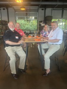 Four people are sitting together at a high table in a brewery-style venue. Each person has a name tag, and there are drinks and napkins on the table. The background features barrels and a window view of greenery outside.