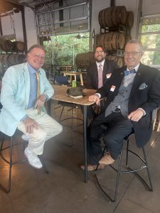 Three individuals are seated at a high table in a brewery-like setting with barrels in the background. They are all dressed in formal attire, including suits and ties. One person is wearing a light blue jacket, another a dark suit with a bow tie, and the third a dark suit with a regular tie.