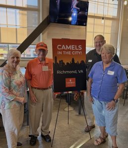 A group of four people stands smiling next to a sign that reads "Camels in the City! Richmond, VA." The setting is an indoor venue with large windows in the background, revealing an urban scene outside.