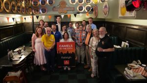 A group of people are gathered in a cozy restaurant with dark wood paneling and green booths. They are standing and smiling around a sign that reads "Camels in the City! Raleigh." The walls are decorated with vintage advertisements and memorabilia. Orange and white pennant banners hang above the group. The atmosphere is warm and friendly.
