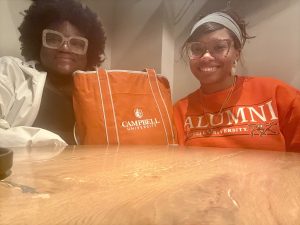 Two people are seated at a wooden table, smiling. One person holds an orange Campbell University tote bag. The other person is wearing a Campbell University Alumni T-shirt. Both are wearing glasses.