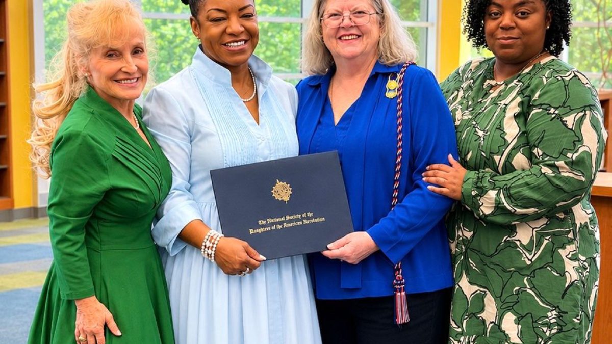 Four people stand together indoors, smiling. One person holds a certificate from the National Society of the Daughters of the American Revolution. They are dressed in formal attire against a background of large windows and bookshelves.