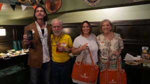 Four people stand together in a room decorated with festive flags. They are each holding items, including insulated cups and orange tote bags labeled "Campbell". A table with food is visible in the background.
