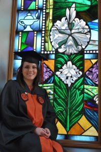 Lauren Banker ’14 wears her Campbell University cap and gown while posing beside a colorful stained-glass window in Butler Chapel.