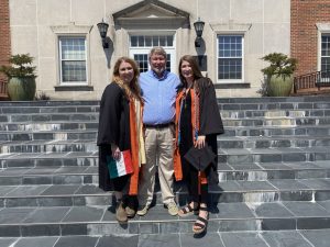 Two Campbell graduates in academic regalia pose with a man on the steps outside a campus building.