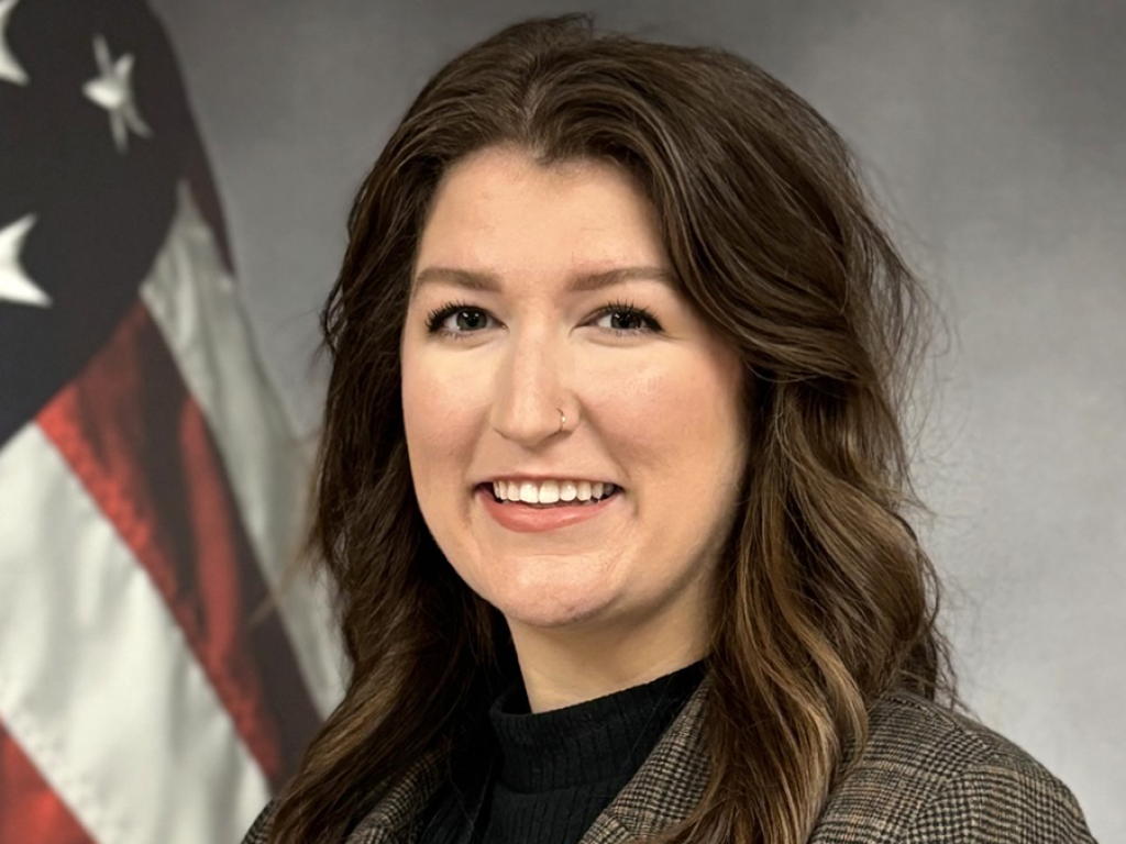 Hannah Byrd Collins poses, smiling for a headshot. An American flag is in the background.