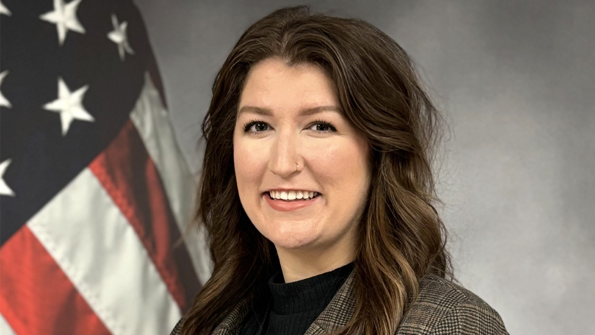 Hannah Byrd Collins poses, smiling for a headshot. An American flag is in the background.