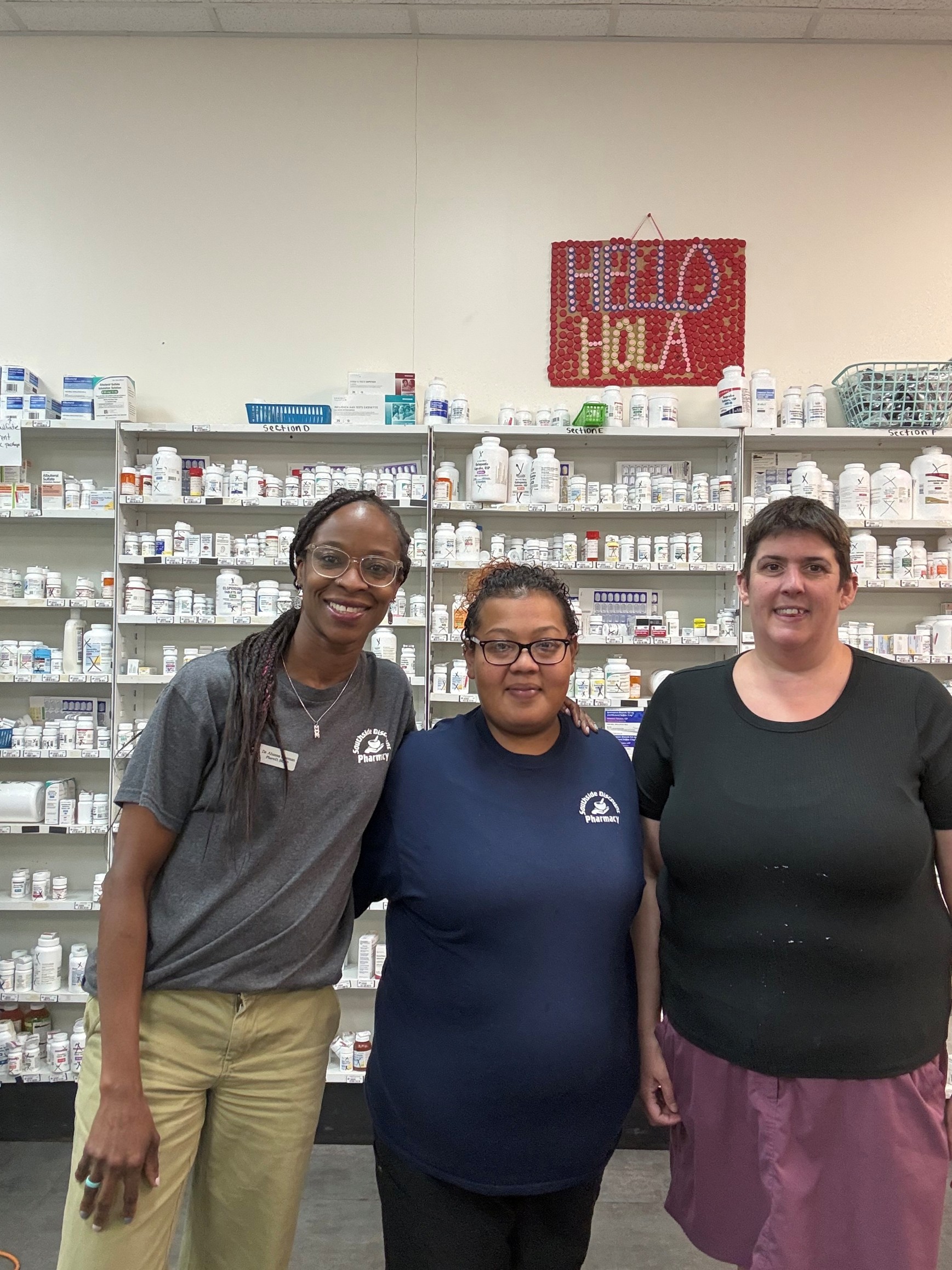 Three females standing in a pharmacy with rows of medication in the background.